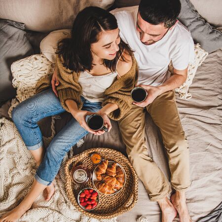 Young smiling happy couple lying on pastel colored blankets barefoot, having breakfast in bed with fresh croissants, strawberries and tea and hugging, top view, square crop. Comfort living conceptの写真素材