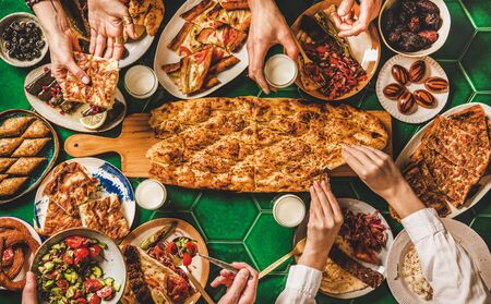 Muslim Ramadan iftar family dinner. Flat-lay of people eating over Middle East food table. Dates, dolma, kebab, flatbread, pide, borek, sweets, ayran drink, top view. Ramazan fasting Turkish cuisineの写真素材