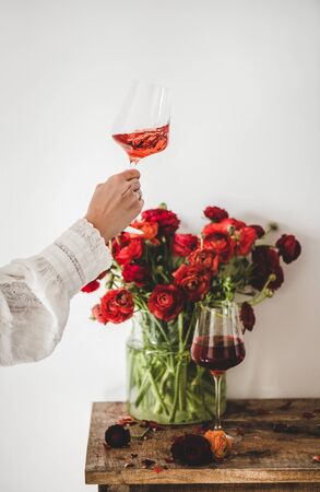 Womans hand with rose wine in glass and red spring blossom flowers bouquet on rustic wooden table over white wall background, selective focus. Wine shop, bar, wine tasting, wine list conceptの写真素材