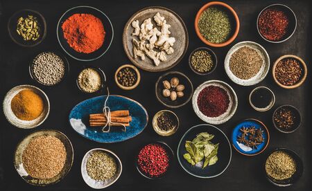 Flat-lay of spices in bowls and plates over dark background, top view. Black pepper, allspice, cloves, thyme, cumin, sesame, star anise, ginger, cinnamon, cardamom, mahlep, smoked salt, rosemary, bayの写真素材