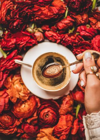 Flat-lay of womans hand holding spoon of black espresso coffee over white porcelain cup and red ranunculus flower background, top view. Seasonal spring or summer coffee break conceptの写真素材