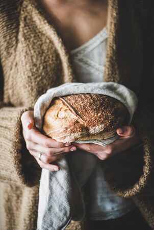 Young woman holding freshly baked healthy wheat Swedish bread round loaf in tpwel in hands,...