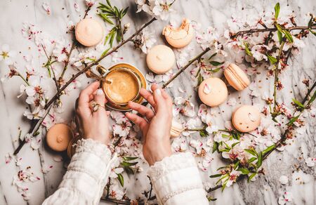 Flat-lay of womans hands over cup of hot espresso coffee, sweet macaron cookies, white spring blossom flowers and white marble background, top viewの写真素材