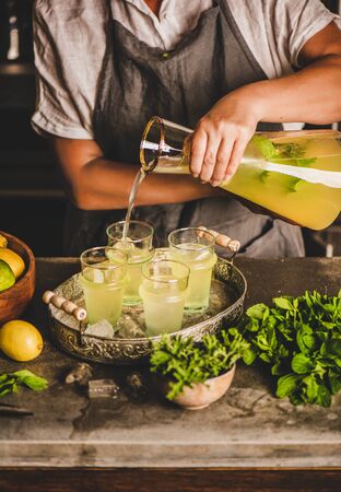 Hands of woman making fresh homemade lemonade over marble kitchen counter. Woman pouring cold lemonade with mint and lemon into glasses with ice in rustic kitchen interiorの写真素材