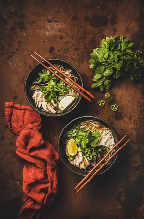 Asian cuisine lunch. Flat-lay of Vietnamese rice noodle chicken soup Pho Ga with cilantro, soy sprouts, fresh greens, lime in black bowls with chopsticks over dark background, top view. Healthy foodの写真素材