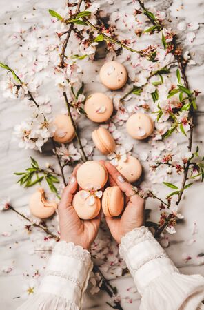 Flat-lay of womans hands holding sweet macaron cookies and white spring blossom flowers and branches over white marble background, top viewの写真素材