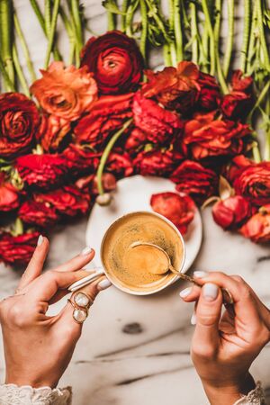 Flat-lay of womans hand holding cup of black espresso coffee over red ranunculus flowers and marble table background, top view. Seasonal spring or summer coffee break conceptの写真素材