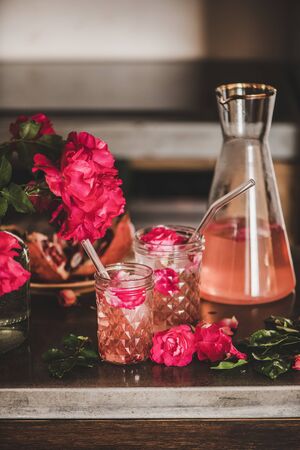 Summer refreshing cold beverage drink. Rose lemonade with ice in glasses and jug and pink rose petals and flowers over concrete kitchen counterの写真素材