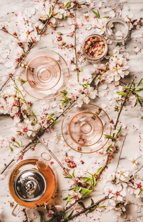 Flat-lay of hot black tea in teapot, glass cups, dry rose flowers in jar and spring blossom almond tree branches over white marble table background, top viewの写真素材