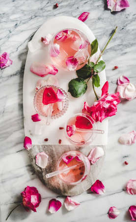 Summer refreshing cold beverage drink. Flat-lay of rose lemonade with ice in glasses and pink rose petals on oval serving board over grey marble table background, top viewの写真素材