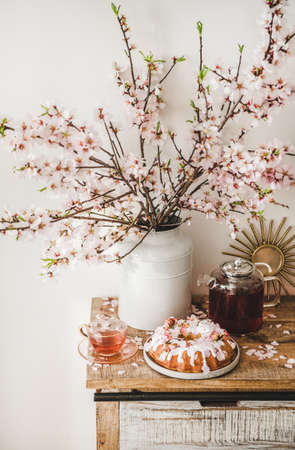 Spring or summer tea and cake setting. Rose and almond gluten-free bundt cake with rose flowers and black tea on rustic wooden cupboard under almond blossom branches in vase, white wall backgroundの写真素材