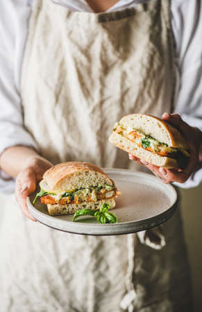 Woman in beige linen apron holding plate with fresh fried fish sandwich with tartare sauce, lemon and arugula cut in halves, selective focus. Healthy easy breakfast ideasの写真素材