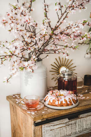 Spring or summer tea and cake setting. Rose and almond gluten-free bundt cake with rose flowers and black tea on rustic wooden cupboard under blooming branches in vase, white wall backgroundの写真素材