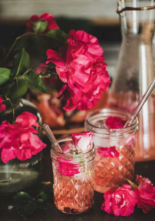 Summer refreshing cold beverage drink. Rose lemonade with ice in glasses and jug and pink rose petals and flowers over concrete kitchen counter, close-upの写真素材