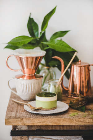 Coffee and dessert setup. Green matcha cheesecake and brewed black coffee in copper pot on rustic wooden kitchen counter, white wall background, close-up. Healthy, vegan, vegetarian, low calorie foodの写真素材