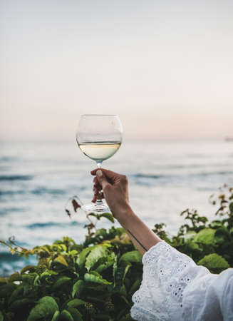 Womans hand in white lace dress sleeve holding glass of white wine with beautiful sunset colors and sea horizon at background. Summer picnic and refreshment drink conceptの写真素材