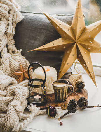 Christmas festive holiday mood composition with coffee in rustic cup, candles and decorative toys on wooden board over white windowsill and woolen blanket at background. Christmas greeting cardの写真素材