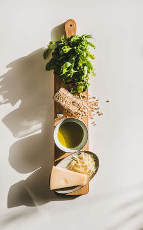 Ingredients for cooking Italian Pesto sauce. Flat-lay of fresh basil leaves, olive oil, pine nuts, garlic, Parmesan cheese on rustic board over white background, top view, vertical compositionの写真素材