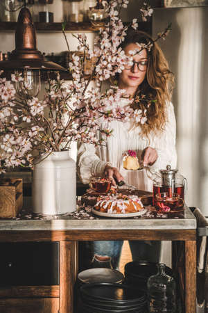 Spring or summer tea and cake setting. Young blond woman serving rose and almond gluten-free bundt cake with rose flowers on concrete kitchen counter with black tea and blooming branches in vaseの写真素材