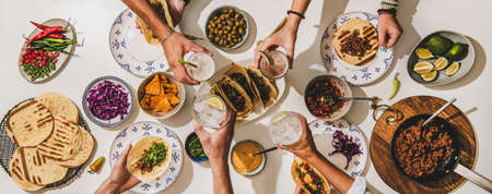 Friends having Mexican Taco dinner. Flat-lay of beef tacos, tomato salsa, tortillas, beer, snacks and peoples hands clinking glasses over white table, top view. Mexican cuisine, comfort food conceptの写真素材