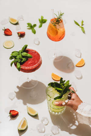Popular bar summer ice alcoholic cocktails. Gin-tonic, and strawberry basil Margarita in glasses and womans hand holding glass over plain white background with cut fruitsの写真素材