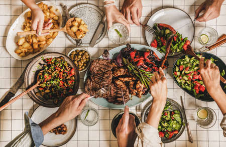 Summer barbeque party. Flat-lay of table with grilled meat, vegetables, salad, roasted potato and peoples hands with cutlery over white tablecloth, top view. Family gathering, comfort food conceptの写真素材