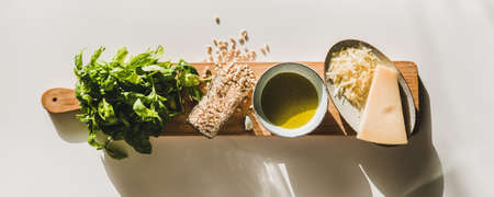 Ingredients for cooking Italian Pesto sauce. Flat-lay of fresh basil leaves, olive oil, pine nuts, garlic, Parmesan cheese on wooden board over white background, top view, horizontal compositionの写真素材