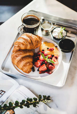 French breakfast cafe table setting. French breakfast with croissant, fresh berries, peach, cream cheese, jam and black coffee in sunlight over white marble table background. Comfort food conceptの写真素材