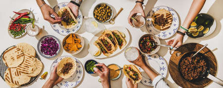 Friends having Mexican Taco dinner. Flat-lay of beef tacos, tomato salsa, tortillas, beer, snacks and peoples hands over white table, top view. Mexican cuisine, gathering, feast, comfort food conceptの写真素材
