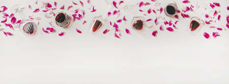 Red wine in glasses. Flat-lay of wine glasses with red wine and pink flowers petals over plain white background, top view, copy space. Wine tasting, winery, bar or Beaujolais Nouveau conceptの写真素材
