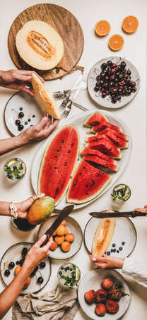 Summer tropical fruit party table. Flat-lay of lunch with berries, watermelon, lemonade and people cutting and holding fruits over white background, top view. Clean eating, fruiterian food conceptの写真素材