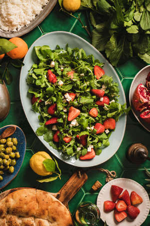 Turkish Mediterranean dinner table setting. Flat-lay of arugula, soft cheese and strawberry salad, rice pilav, flatbread and olives over oriental green tiled table background, top view. Turkish regional Aegean cuisineの写真素材
