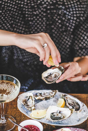 Hands of woman squeezing lemon juice to fresh Irish oysters over ice in plate and glass of champagne in fish restaurant, selective focus. Seafood, French cuisine, fine dining conceptの写真素材