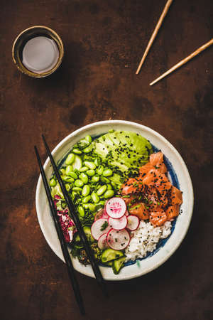 Healthy dinner. Flat-lay of salmon poke bowl or sushi bowl with vegetables, greens, sushi rice, soy sauce and bamboo sticks over dark rusty table background, top view. Traditional Hawaiian cuisineの写真素材