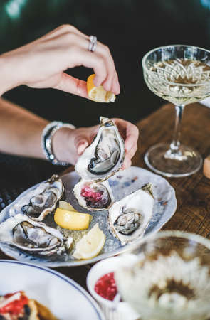 Hands of woman squeezing lemon juice to Irish oysters in plate with ice over glass of champagne in fish restaurant at background, selective focus. Seafood, French cuisine, fine dining conceptの写真素材