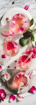 Summer refreshing cold beverage drink. Flat-lay of rose lemonade with ice cubes in glasses and rose flower petals on oval serving board over grey marble table background, top view, wide compositionの写真素材