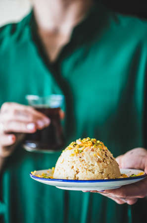 Turkish traditional dessert semolina helva. Womans hand holding plate with sweet semolina helva with pistachio and glass of Turkish tea, selective focus. Turkish cuisineの写真素材