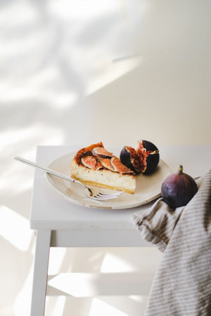 Slice of homemade gluten free fig cheesecake with fresh seasonal figs on white plate over white wall with sun shadows at background. Healthy dessert, artisan sweet food conceptの写真素材