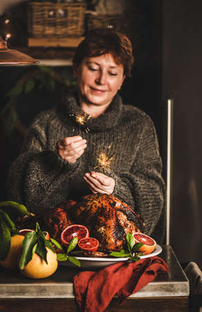 Smiling woman in grey sweater burning sparkles over whole roasted turkey or chicken in oranges for festive holiday table setting. Christmas and New Year holiday celebration and traditional foodの写真素材