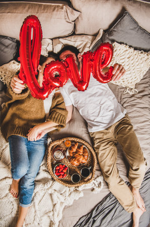 Young happy couple lying on pastel colored blankets barefoot in bed, having breakfast with fresh croissants, strawberries and tea, holding hands and sign love, top view. Saint Valentines day conceptの写真素材