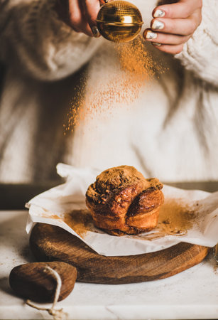 Female hands sprinkling ground cinnamon powder over sweet warm cinnamon caramel bun on rustic wooden board, selective focus. Winter dessert, comfort food conceptの写真素材