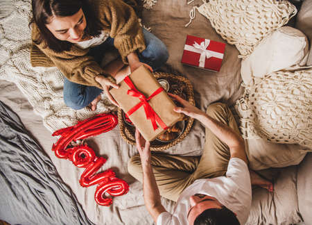 Young happy couple sitting on blankets in bed at home and giving presents in boxes over fruits and sign love on bed, top view. Saint Valentines day conceptの写真素材