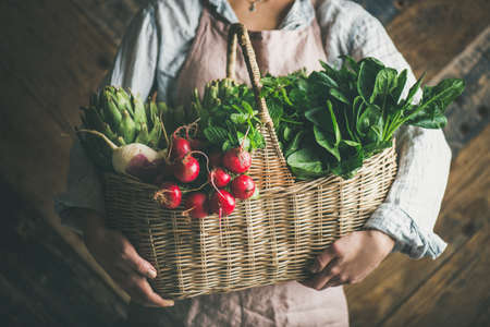 Woman farmer holding basket of fresh organic garden vegetables and greens in hands, rustic wooden wall at backgroundの写真素材