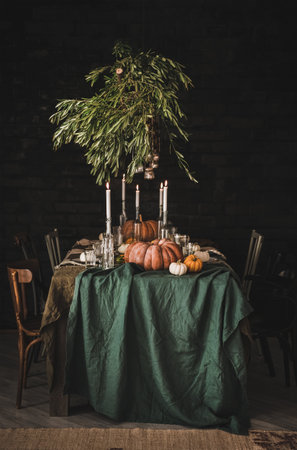 Stylish rustic Thanksgiving Day party table setting. Solid table with green linen tablecloth, dinnerware, glassware and cutlery decorated with pumpkins, glass candle holders and olive tree branchesの写真素材