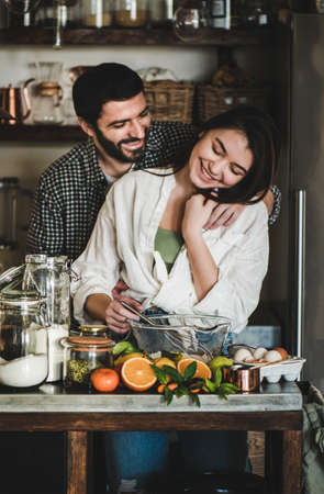 Young loving couple cooking together in kitchen. Young caucasian man and brunette woman hugging, flirting, laughing and making dough for baking citrus cake. Valentines Day, home hobbies conceptの写真素材