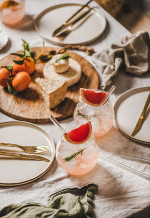 Cocktail party table setting. Grapefruit rosemary margaritas in glass, cheese fruit board, dinnerware, cutlery on linen tablecloth. Romantic table setting for celebration, stay at home during lockdownの写真素材