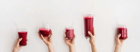 Human hands holding glasses with purple beet pomegranate fresh homemade smoothie with glass straws, white background, wide composition. Detox, dieting, weight loss, healthy lifestyle conceptの写真素材