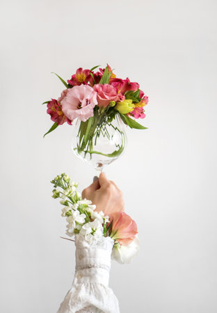 Wine glass full of fresh pink, red and white Spring flowers in hand of woman in white blouse, white background. Wine smell creative or Womens Day conceptの写真素材