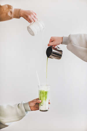 Female hands pouring japanese matcha tea and milk from jug into glass for making green matcha latte for breakfast over light wall background. Coffee beverage drinks and japanese cuisineの写真素材
