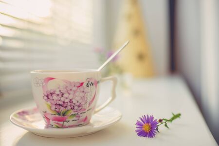 Elegant vintage porcelain cup and spoon on the white windowsill, side view with warm sunlight
